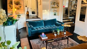 Una sala de estar con un sofá color turquesa, una mesa de centro con libros, una alfombra y una escalera que lleva a una cocina.