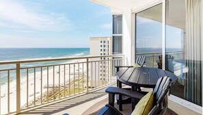 Ein Balkon mit Blick auf Meer und Strand, ein Tisch mit Stühlen und ein großes Fenster mit Vorhängen.