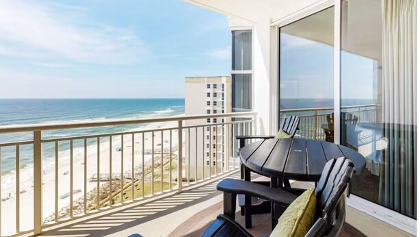 A balcony with a view of the ocean and beach, a table with chairs, and a large window with curtains. - Palacio 803 (Pensacola)