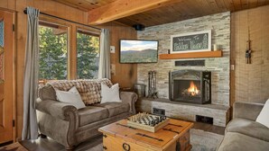 A cozy living room with a fireplace, a sofa, a coffee table with a chess set, and a television. - Hidden Pines - Hot Tub, BBQ & Pool Table (Big Bear Lake)