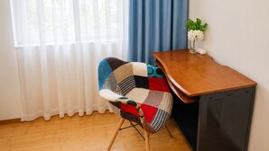 Ein Zimmer mit Schreibtisch aus Holz, einem Sessel mit Patchwork-Kissen, einer kleinen Pflanze und einem Fenster mit durchsichtigen Vorhängen.