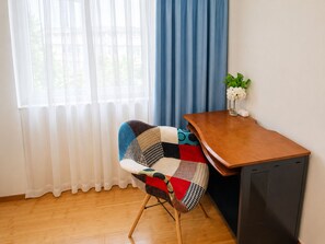 Ett rum med ett skrivbord i trä, en stol med ett lapptäcksöverdrag, en liten växt och ett fönster med tunna, genomskinliga gardiner.