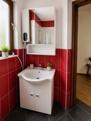 Et badeværelse med en hvid vask, røde fliser på væggene, et spejl over vasken og et vindue med en plante.