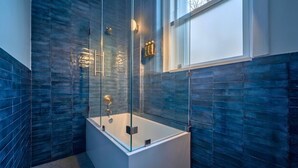 A modern bathroom with a glass-enclosed bathtub, a large window, and a blue-tiled wall.