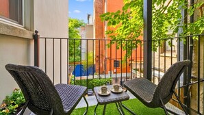 A balcony with two wicker chairs and a small table, a view of a brick building, and a blue fence in the background. - Prime Location 2BDR Loft: Patio, Steps to Subway (New York)