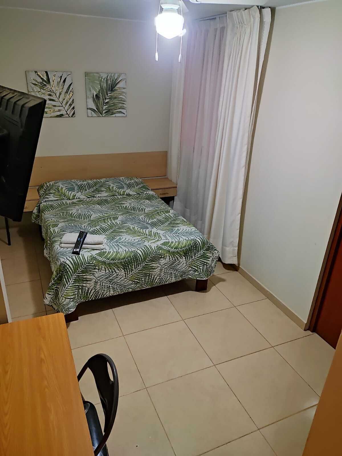 Una habitación pequeña y ordenada con una cama, un escritorio y una silla. Hay dos cuadros colgados en la pared y una lámpara en el techo.