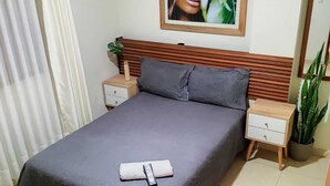 A neatly made bed with a dark bedspread and pillows, a wooden headboard, a bedside table with a lamp, and a potted plant beside the bed. - kallma adventures (Ica)