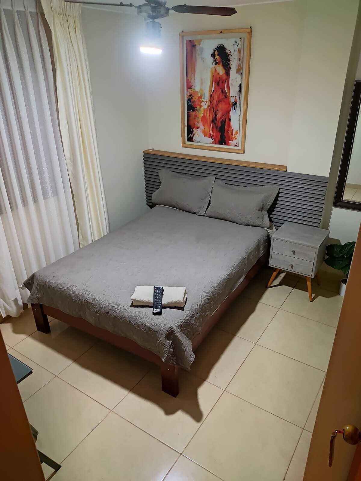 A neatly made bed with a grey bedspread and pillows, a bedside table with a lamp, and a painting on the wall.
