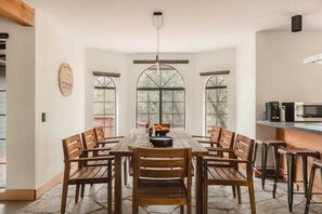 Un espace repas avec une longue table en bois et des chaises, un lustre central et de grandes fenêtres surmontées d’arcs.