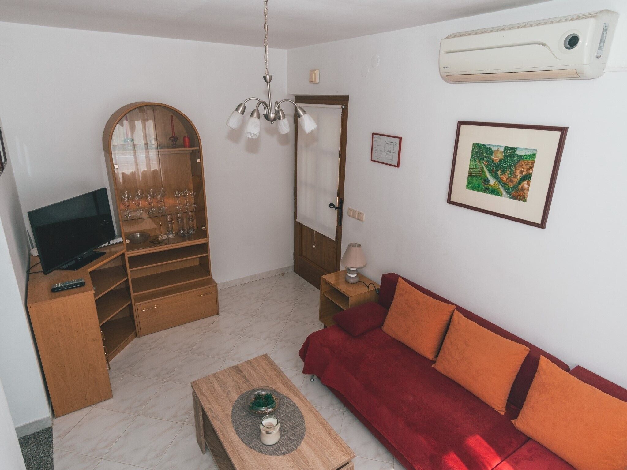 A living room with a TV, wooden furniture, a red sofa, a glass cabinet with wine glasses, and a painting on the wall.