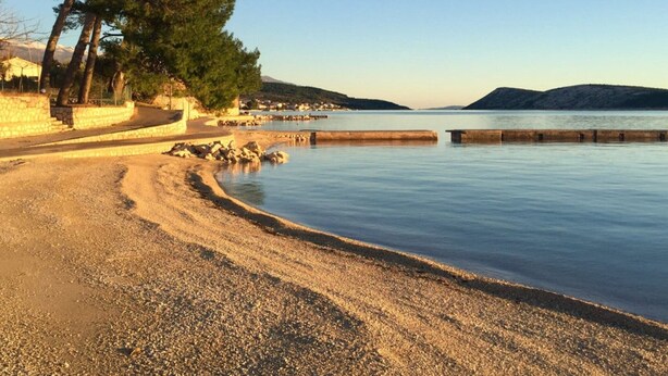A pebbled beach with a calm sea, a stone wall, and a few trees.