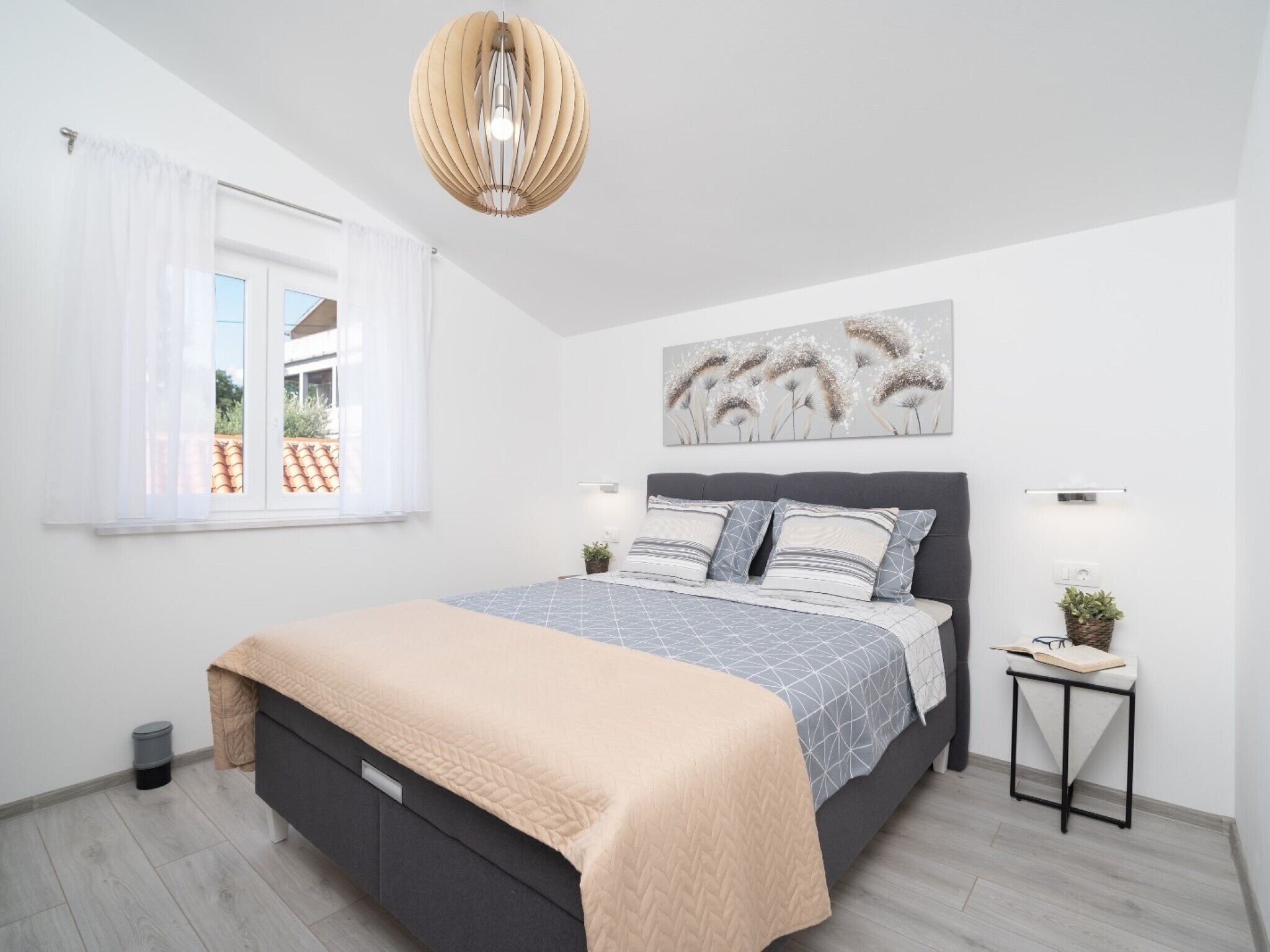 A bedroom with a bed, bedside tables, a hanging light fixture, a window with curtains, and a wall-mounted artwork.