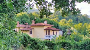 A two-story house with a red-tiled roof, surrounded by lush greenery and trees.