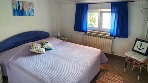 Une chambre à coucher avec un lit, une fenêtre avec des rideaux, un radiateur, une petite table avec une chaise et un tableau accroché au mur.