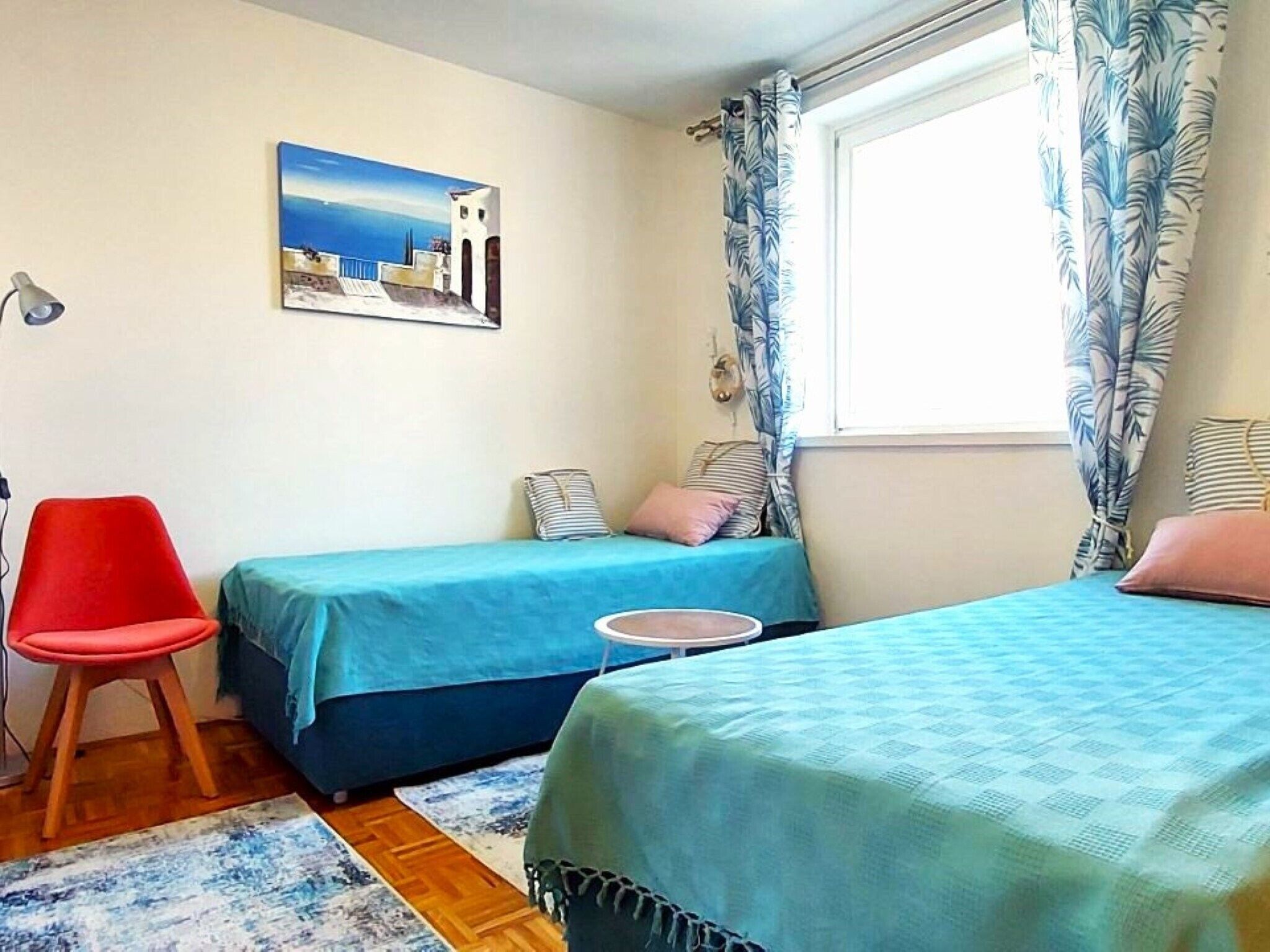 A bedroom with two beds, a red chair, a small round table, and a painting on the wall.