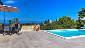 Une piscine avec vue sur la mer et les montagnes, un espace extérieur aménagé avec une table et des chaises, et des plantes en pot.