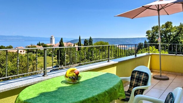 Un balcon avec une table recouverte d’une nappe verte, un saladier rempli de fruits et un grand parasol.