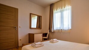 Une chambre d’hôtel avec un lit, une armoire en bois, un bureau, une chaise, un miroir et une fenêtre avec des rideaux.