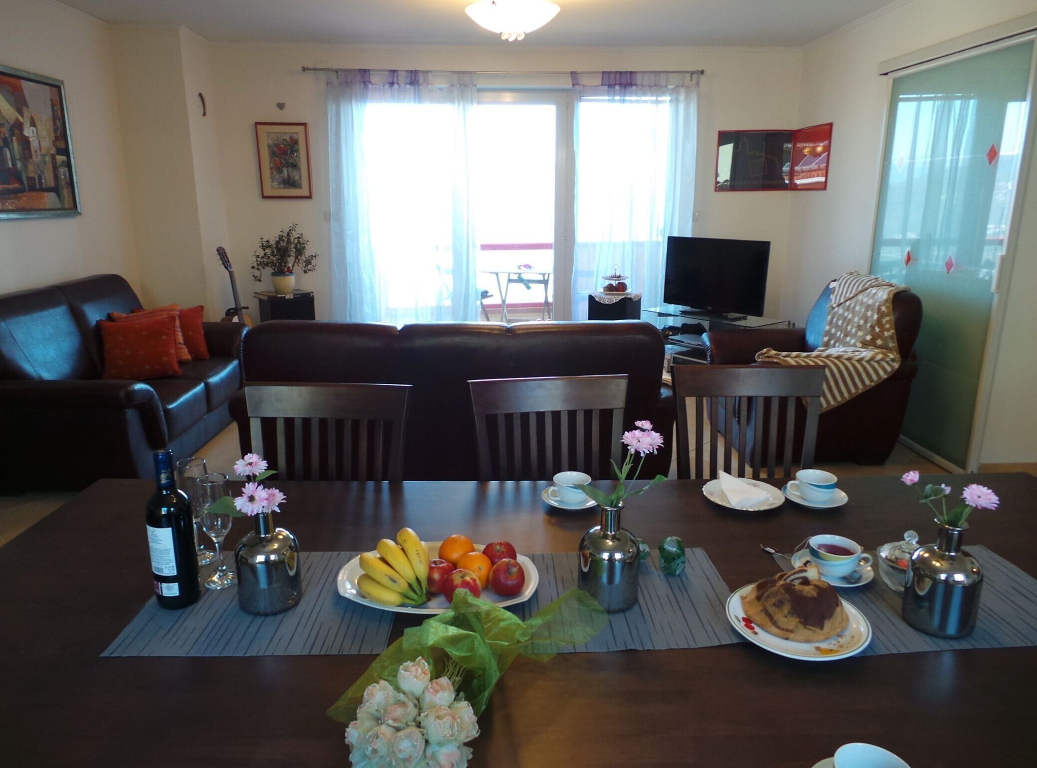 A dining area with a table set for a meal, chairs, a sofa, a television, and a window with curtains.