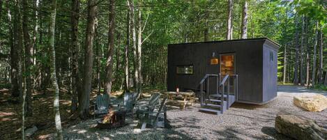 A cabin in a forest with a gravel ground, wooden benches, and a fire pit.