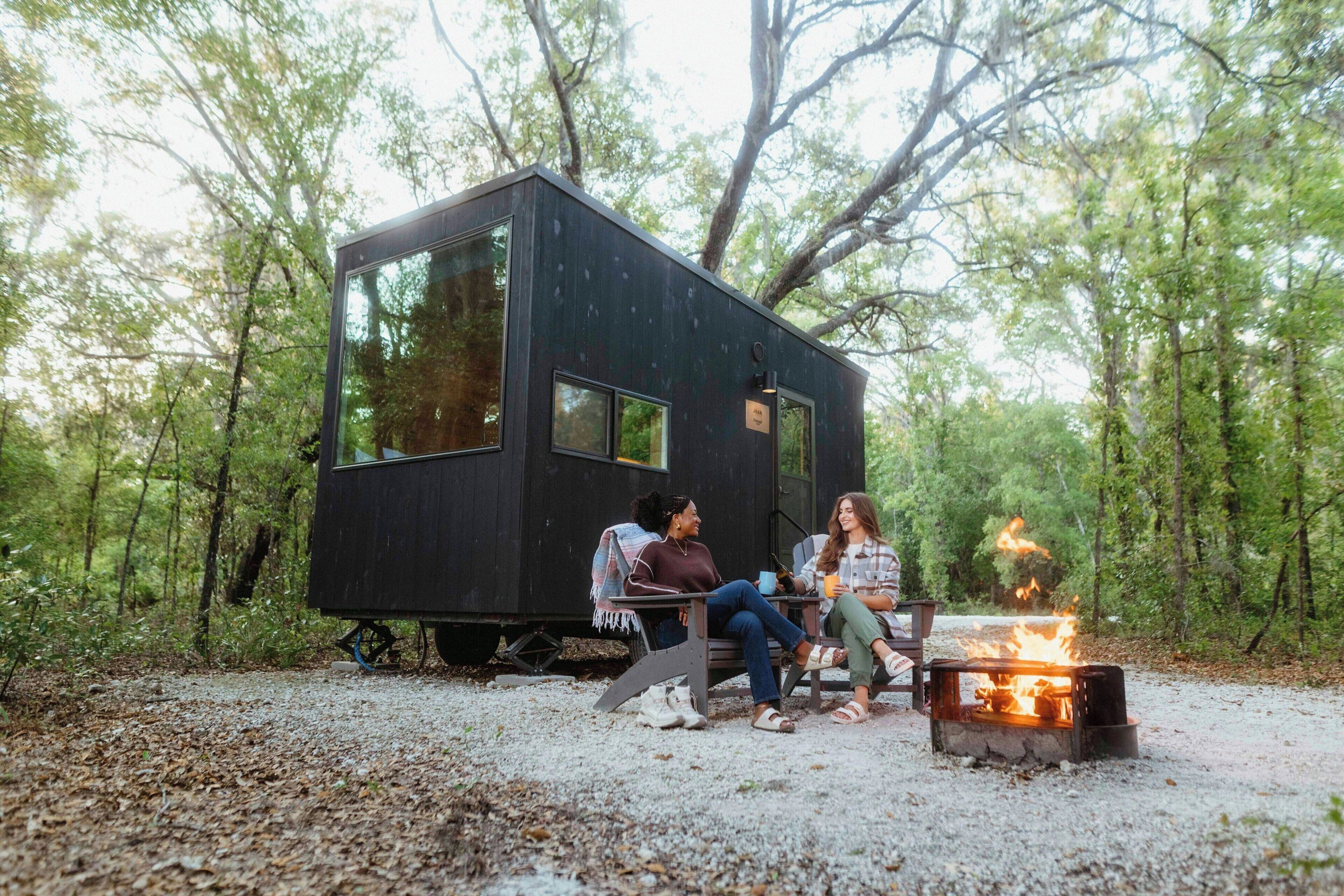 A small, dark-colored cabin with a fire pit in the center, surrounded by a forested area.