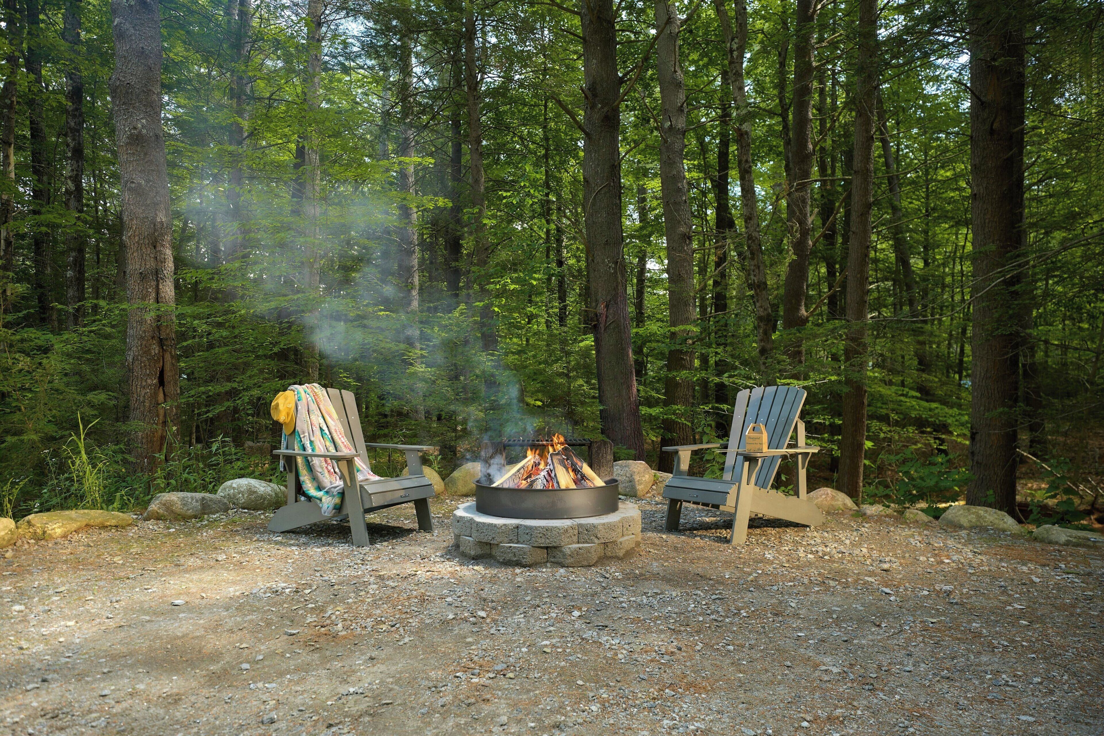 A campsite with two Adirondack chairs, a fire pit, and a forested backdrop.