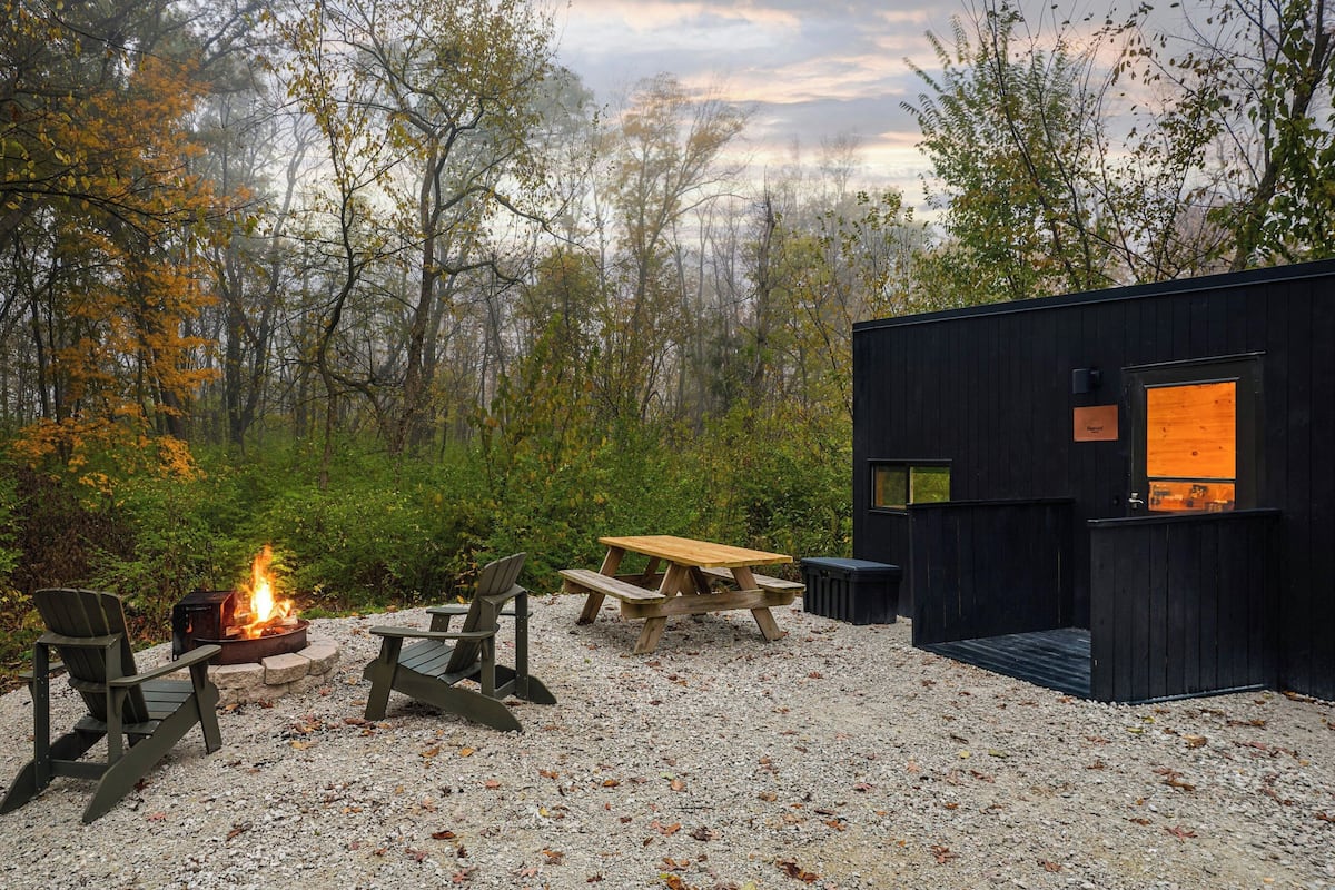 A rustic cabin with a fire pit and seating area, surrounded by trees.