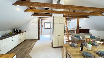 A spacious loft with a high ceiling, exposed wooden beams, a kitchen with modern appliances, a dining area with a wooden table set for six, and a cozy seating area with a sofa and cushions.