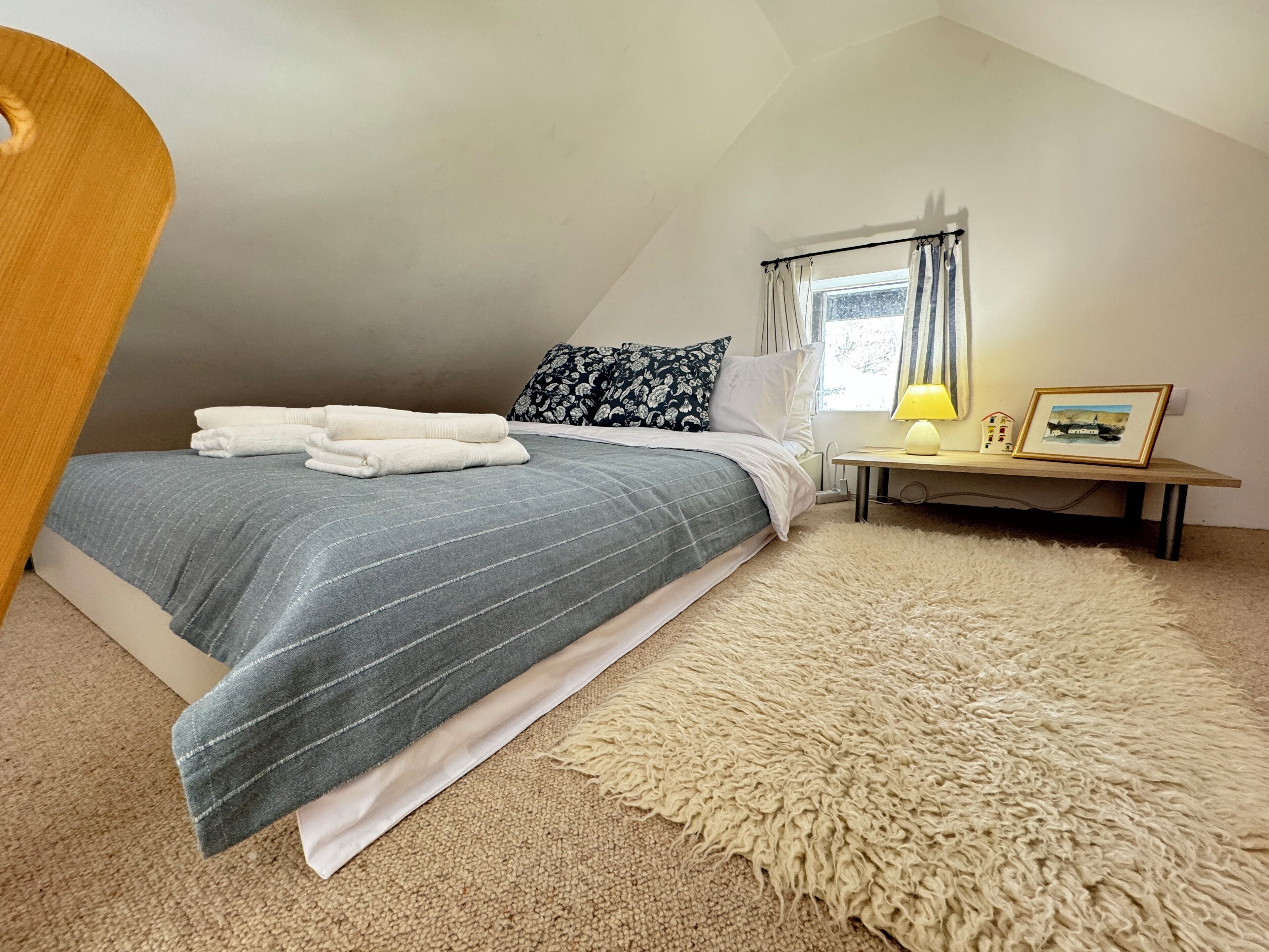 Une chambre à coucher avec un lit, une table de chevet avec une lampe, un tableau encadré et une fenêtre avec des rideaux.