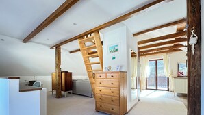 Une chambre spacieuse aménagée en mezzanine, avec un escalier en bois menant à une zone de couchage en hauteur, une grande commode et une vue sur l’extérieur grâce à une porte-fenêtre coulissante.