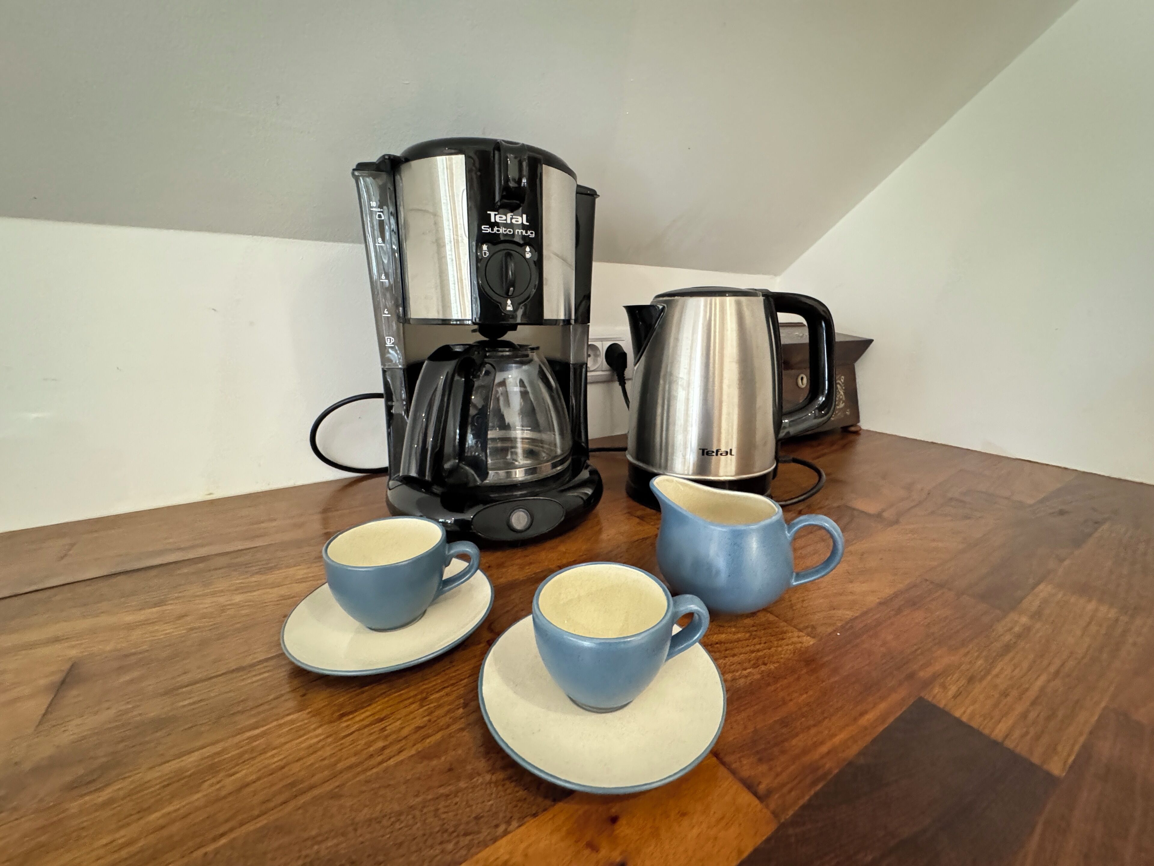 A kitchen with a coffee maker, a kettle, and two cups on a wooden table.