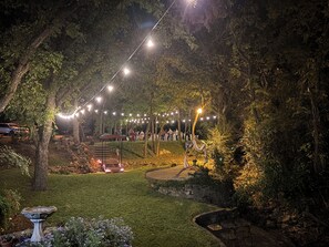 Ein Gartenbereich im Freien mit Lichterketten, einer Steintreppe und einem Essbereich mit Tischen und Stühlen.
