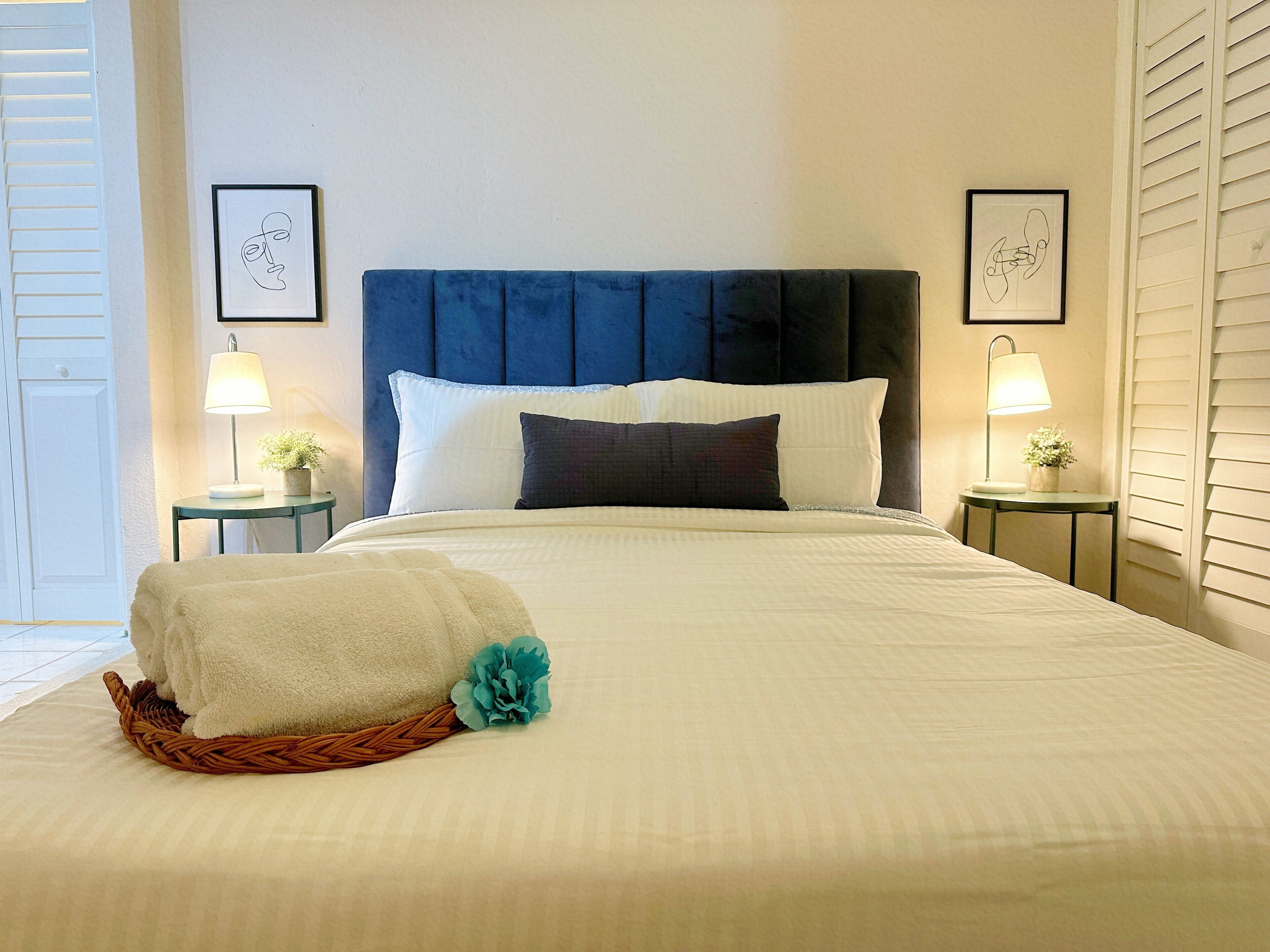 Una cama bien hecha con cabecera azul, almohadas blancas y oscuras, y una colcha blanca. Hay dos mesitas de noche con lámparas y pequeñas macetas.