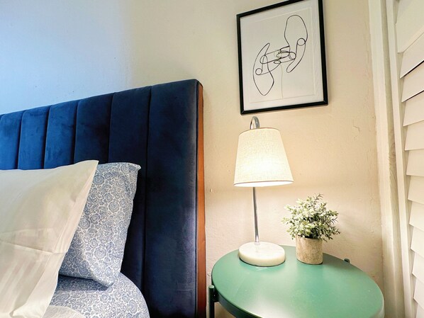A bedroom with a blue upholstered headboard, a bedside lamp, a framed abstract artwork, and a small potted plant on a green side table. - Jetsetter's Retreat (Carolina)