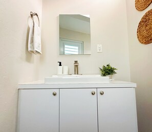 A bathroom with a white vanity, a mirror, a sink with a faucet, a towel hanging on the wall, and a small plant on the counter. - Jetsetter's Retreat (Carolina)