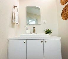 Un baño con un tocador blanco, un espejo, un lavabo con grifo, una toalla colgada en la pared y una planta pequeña en la encimera.