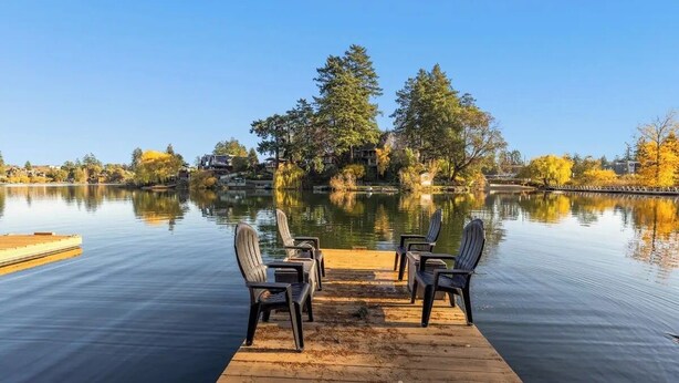 A wooden dock with four Adirondack chairs facing a calm lake, surrounded by trees and buildings in the background. - Lakefront Escape & Water Access (Victoria)