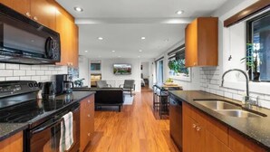 A modern kitchen with wooden cabinets, a black countertop, and a stainless steel sink. There is a dining area with a black sofa and a table. - Lakefront Escape & Water Access (Victoria)
