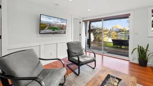 A modern living room with a large TV mounted on the wall, two grey armchairs, a wooden coffee table, and a large sliding glass door leading to an outdoor area. - Lakefront Escape & Water Access (Victoria)