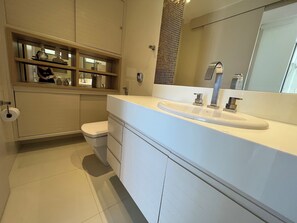 A modern bathroom with a white sink, a toilet, and a mirror above the sink. There are shelves with toiletries and a patterned wall behind the mirror. - Toque a Campainha 402 Casal  (Cabo Frio)