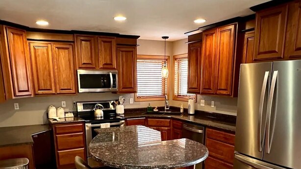 A kitchen with wooden cabinets, a stainless steel refrigerator, a microwave, and a granite countertop island with two bar stools. - Modern Comforts - Stay/Work/Play (Casper)