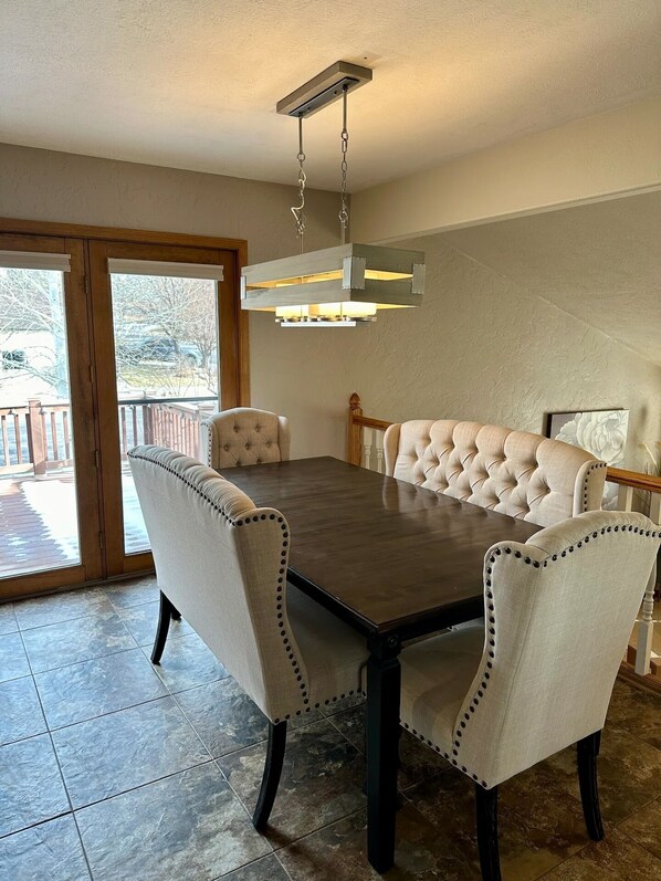 A dining area with a dark wooden table and tufted chairs, a hanging light fixture, and a view through a glass door to an outdoor deck. - Modern Comforts - Stay/Work/Play (Casper)