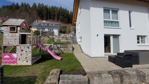 Ein modernes, zweistöckiges Haus mit Natursteinfassade, ein hölzernes Spielhaus inklusive Rutsche und eine gepflasterte Terrasse.