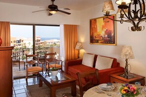 A hotel room with a balcony, a sofa, a dining table set, and a painting on the wall.
