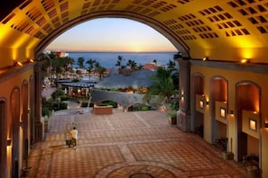 A hotel corridor with a view of the ocean at sunset, featuring a stone bridge and outdoor seating area.