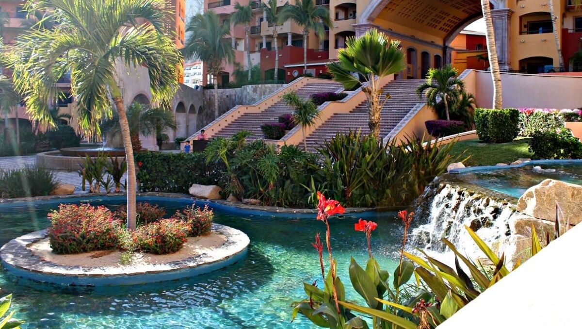 A hotel courtyard with a pool, palm trees, a waterfall, and a staircase leading to an upper level.