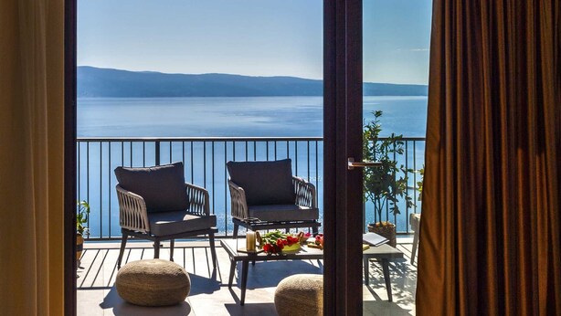 A balcony with two wicker chairs, a small table with a vase of flowers, and a view of a calm body of water and distant mountains. - Charming 3BR Villa for 6, With Heated Pool with Waterfall (Omiš)