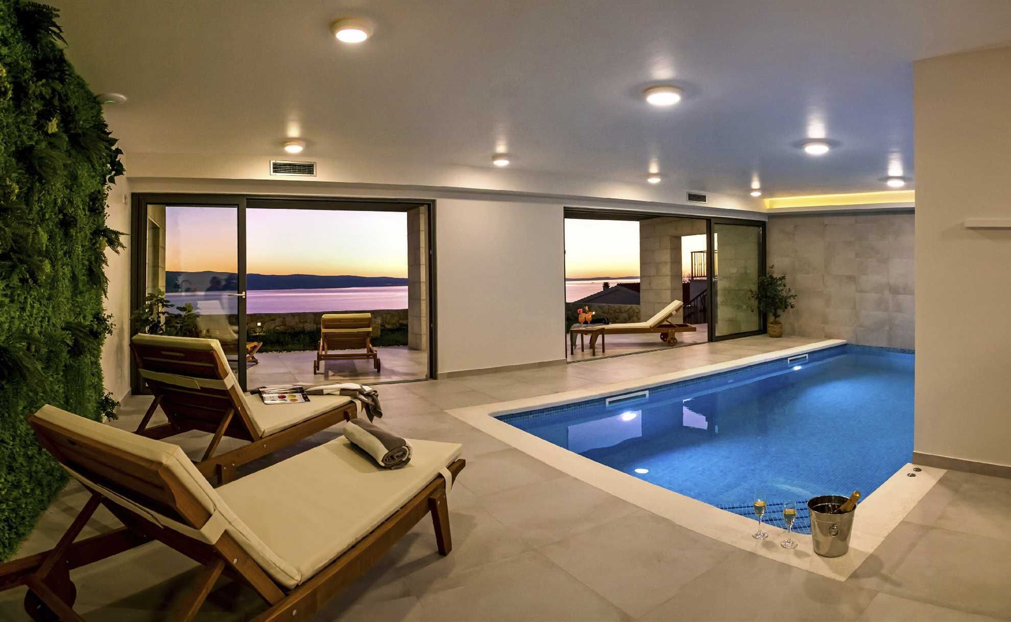 A modern indoor swimming pool with lounge chairs and a view of the sunset.