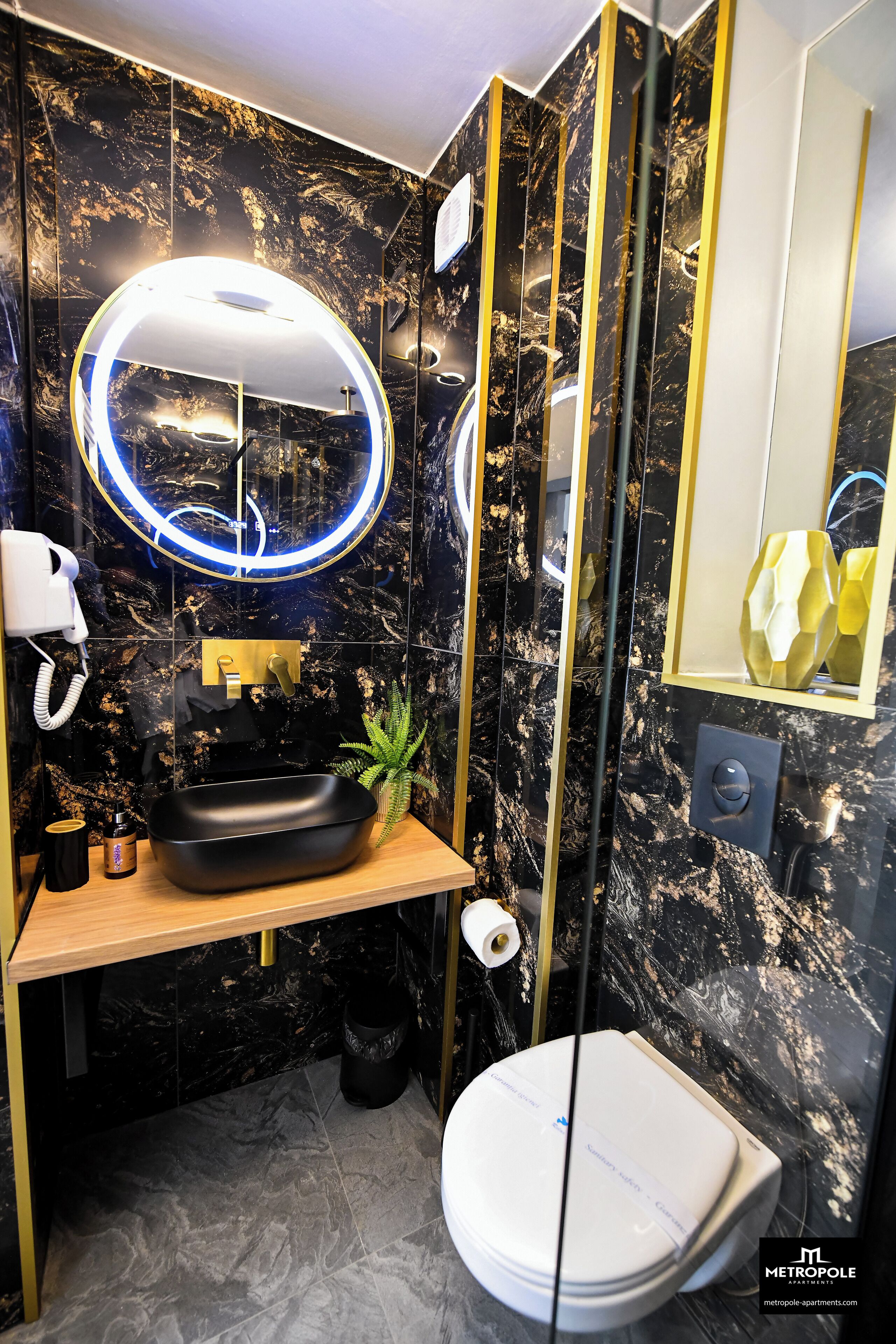 Ein Badezimmer mit schwarzem Marmor an der Wand, einem Holzbadschränkchen, einem runden Spiegel, einer weißen Toilette und einem kleinen Topfpflanzen.