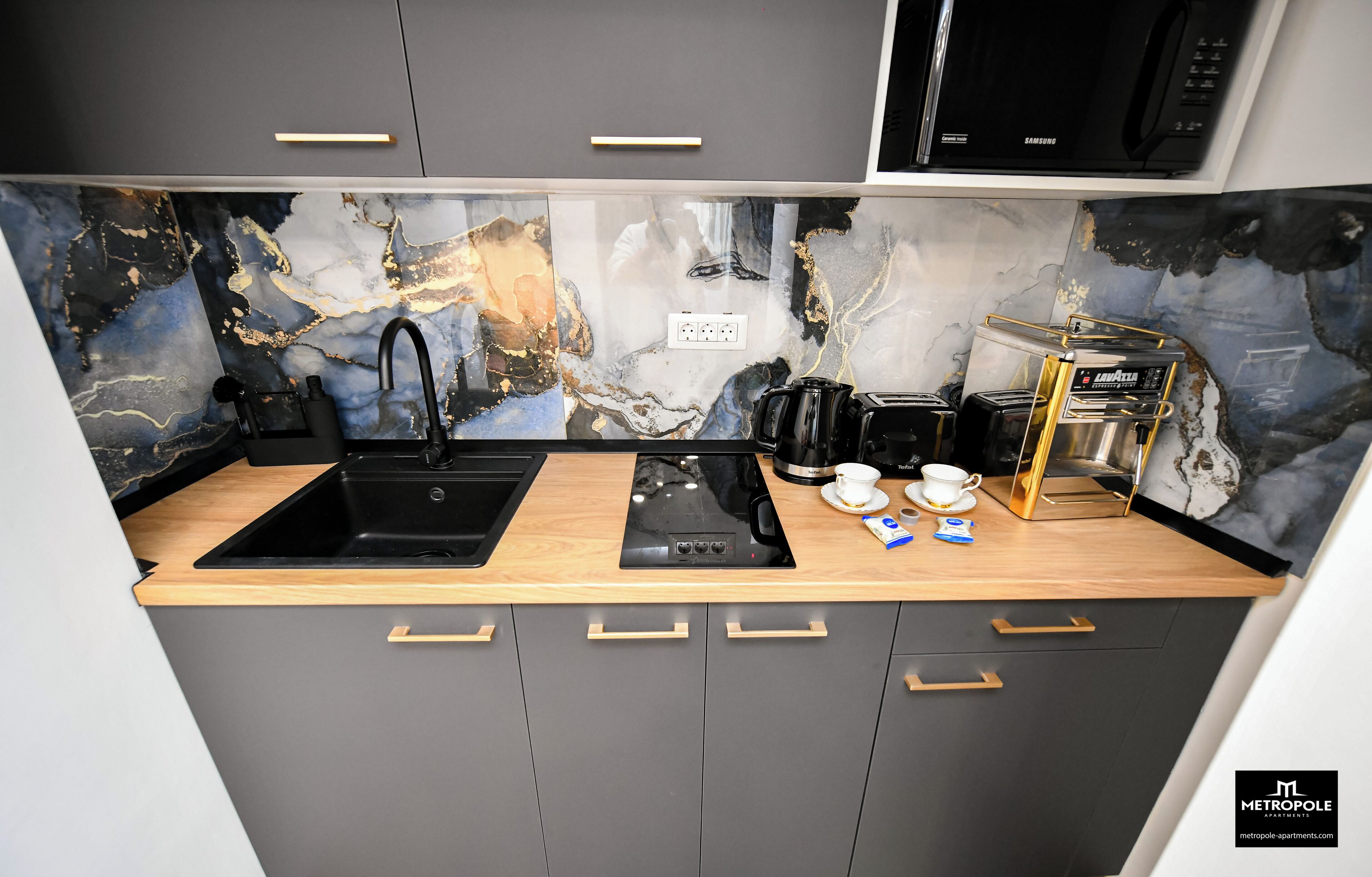 A modern kitchen with a black sink, a built-in stove, and a microwave. There are cabinets with gold handles and a marble backsplash.